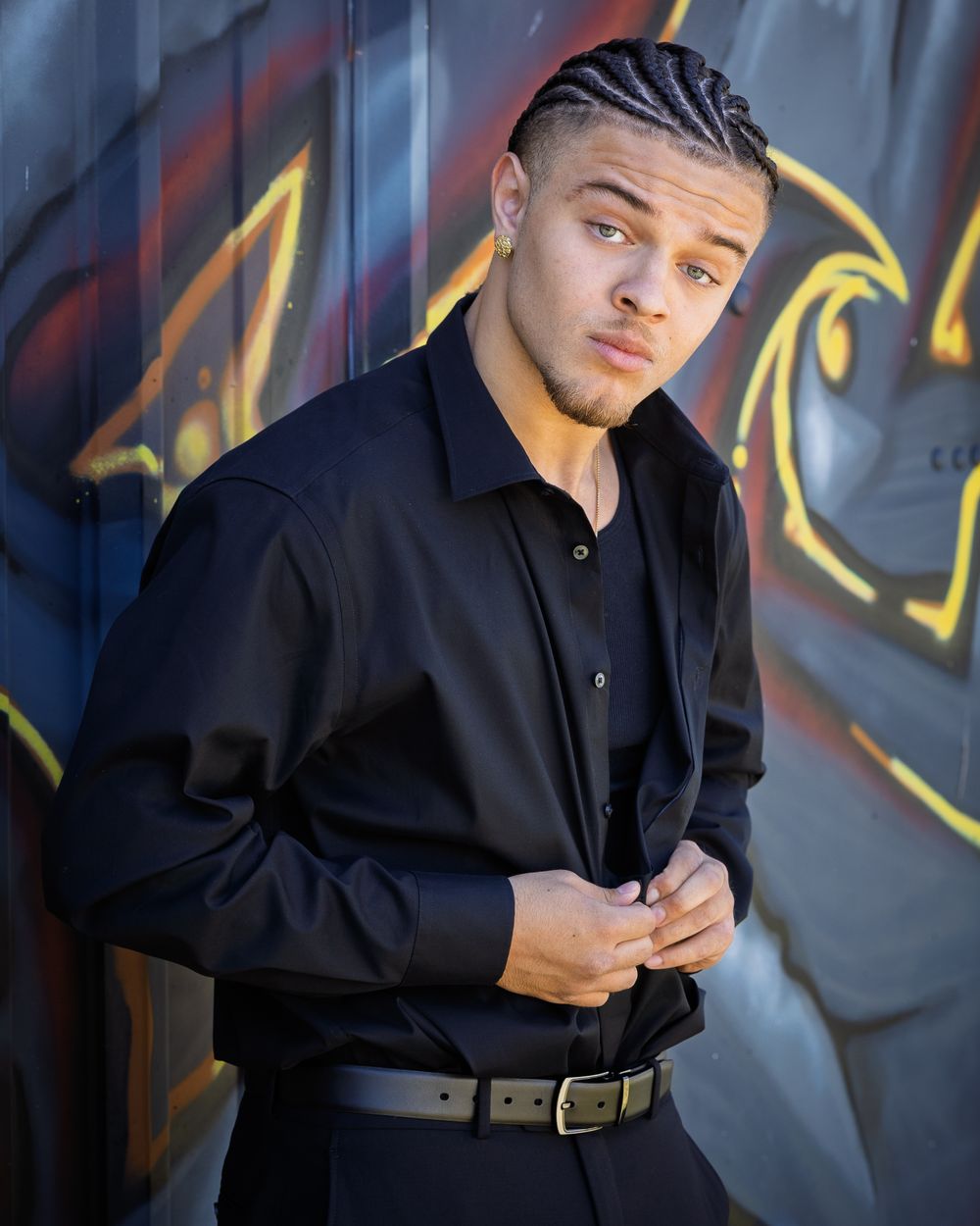 Young man with braids in black shirt and belt poses confidently against colorful graffiti wall.