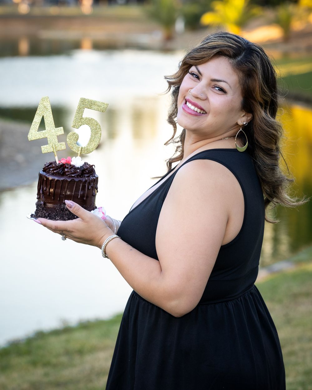 Woman in black dress smiling and holding a chocolate birthday cake with gold number 45 topper outdoors near a pond.