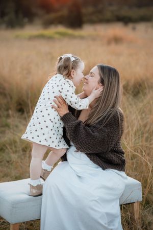 A mother and toddler daughter share a tender nose-to-nose moment outdoors, sitting on a bench in an autumn field.