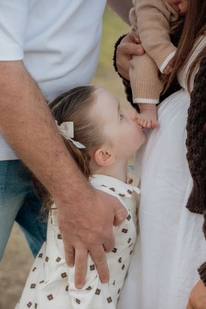A young girl in a floral dress kisses a baby's hand while standing between her parents outdoors.