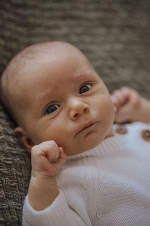 A baby wearing a white knit sweater lies on a textured gray surface looking up at the camera.