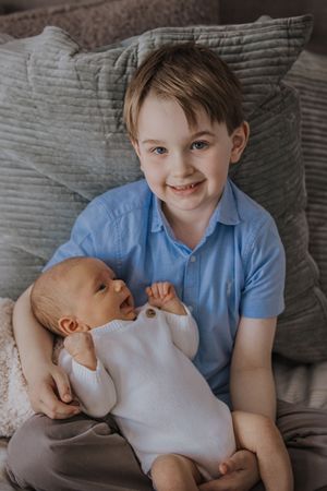 A young child in a blue shirt holds and gazes lovingly at a newborn baby in a white onesie on a gray couch.