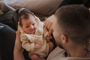 A smiling baby wearing a cream-colored sweater lies on a couch while being lovingly engaged with.
