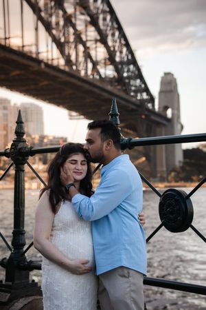 Romantic couple embraces by the harbor railing with iconic Sydney Harbour Bridge in the background at sunset.