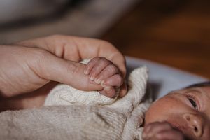 A tiny newborn baby's hand grasps an adult finger while wrapped in a soft cream-colored blanket.