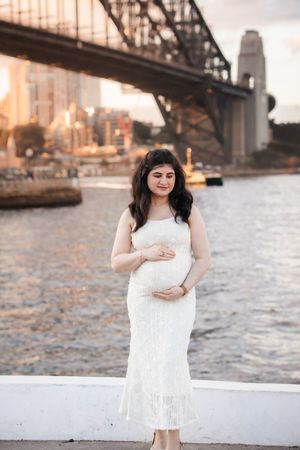 A maternity photoshoot at sunset by the waterfront with a bridge in the background.