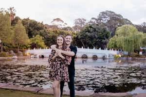 A romantic couple embraces by a tranquil lily pond in a scenic garden setting with stone bridge and willow trees.