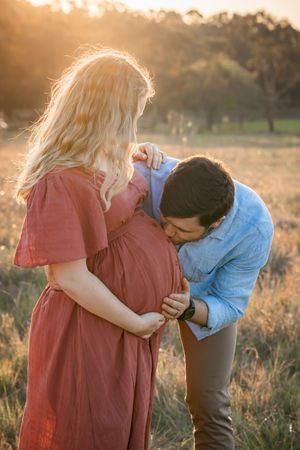 A couple in a field at sunset sharing a tender moment as they look down at her pregnant belly.