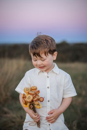 A child in a white shirt holds a bouquet of wildflowers while standing in a field at sunset.