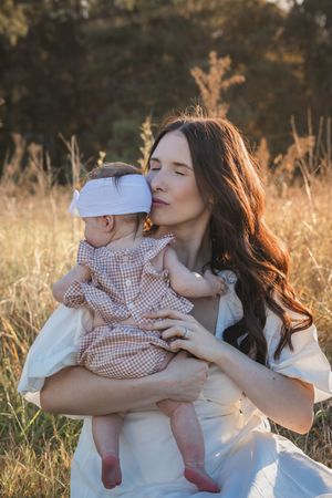 A tender moment captured in a golden field at sunset as a mother in white embraces her baby in gingham dress.