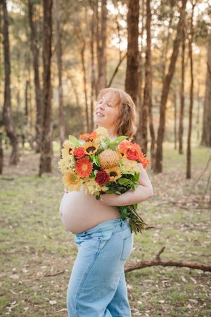Person in overalls embraces colorful sunflower and rose bouquet in sunlit forest setting at golden hour.