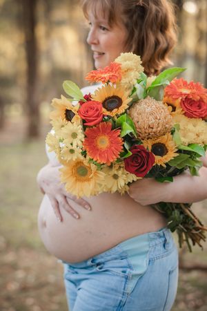 A pregnant woman in jeans holds a vibrant autumn bouquet with sunflowers, coral gerberas, and roses against a blurred background.