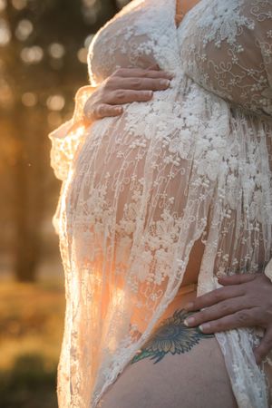 A pregnant woman in a white lace dress cradles her belly during golden hour sunset photography.