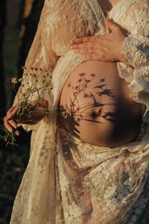A pregnant woman in lace dress holds dried flowers casting shadows on her belly in warm sunlight.
