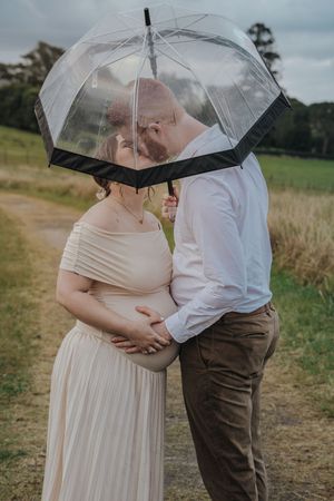 A couple shares an intimate moment under a clear umbrella in a grassy field on an overcast day.