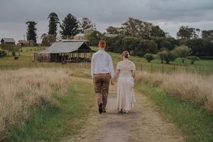 Two people walk hand in hand down a dirt path through a grassy field on an overcast day.