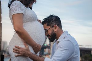 A person in a white shirt kisses a pregnant woman's belly while standing outdoors during a maternity photoshoot.