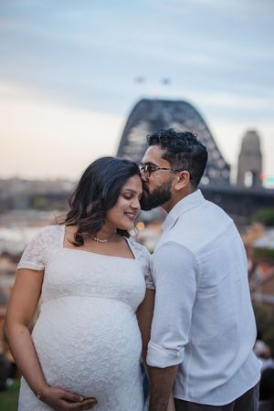 A romantic couple in white outfits share tender moments during an outdoor photoshoot with a city skyline backdrop.