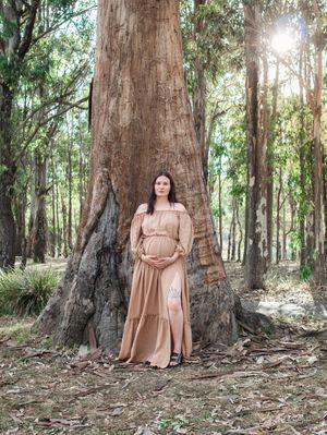 A person in a flowing beige dress poses gracefully against ancient eucalyptus trees in a sunlit forest setting.