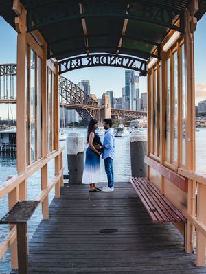 A couple stands together on a covered wooden pier overlooking the Boston harbor and skyline on a sunny day.