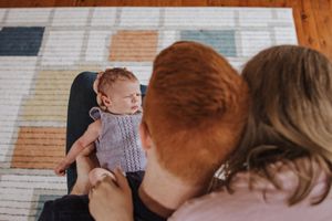 A baby wearing a striped onesie sits with adults on a patterned area rug in a living room setting.