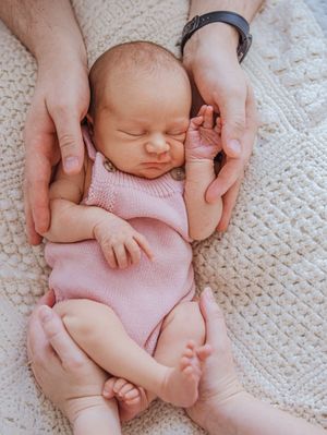 Newborn baby wearing pink sleeper outfit lies peacefully on white textured blanket while gentle hands support them.
