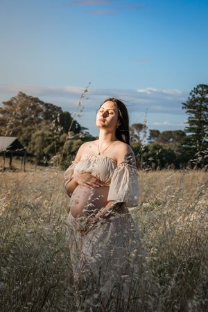 Expectant mother in flowy beige dress poses gracefully in golden field under blue sky during maternity photoshoot.
