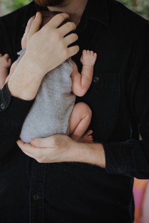 A tiny baby's foot and hand peek out from a gray sweater while being held against someone wearing a black shirt.