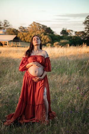 A maternity photoshoot in a rustic field at sunset shows someone in a flowing rust-colored dress cradling their belly.