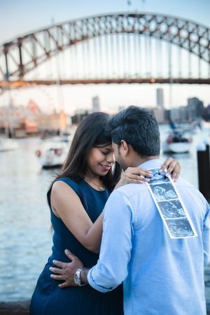 A couple embraces by the Sydney Harbour Bridge with the iconic steel arch structure visible in the background.