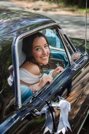 Woman smiling while sitting in vintage black car wearing white dress.