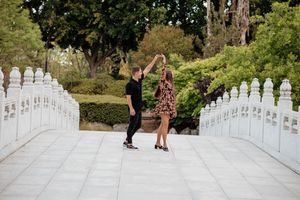 A couple walks along a white stone balustrade staircase surrounded by lush greenery in a garden setting.