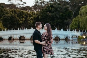 A couple shares a romantic moment by a scenic lake with an ornate white bridge in the background.