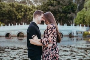 Romantic couple embracing by a lake with stone bridge in background during outdoor photo session.