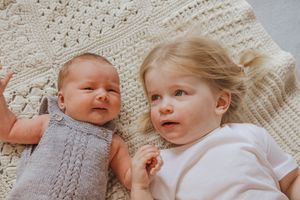 Two babies lying on a cozy knit blanket share a sweet interaction while wearing soft neutral colored outfits.