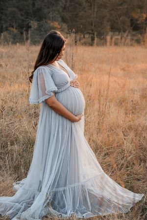 Ethereal maternity photo in a light blue flowing dress standing in golden autumn field at sunset.