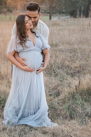 A couple in soft blue attire embraces in a golden field during a romantic maternity photoshoot at sunset.