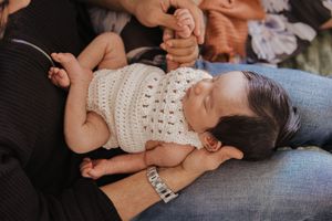 Baby wearing white crocheted top lies on parent's lap during tender moment.