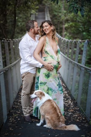 A couple in white and floral dress share romantic moments on a wooden bridge walkway with their fluffy dog.