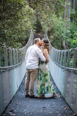 A couple shares a romantic moment on a suspended footbridge surrounded by lush forest greenery.