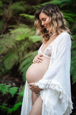 A maternity photo in a forest setting shows an expectant mother wearing a white robe and lace lingerie.