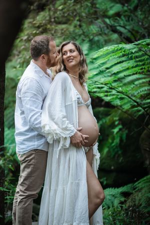 Maternity photoshoot in a lush forest setting with couple wearing white and tan clothing embracing among green ferns.