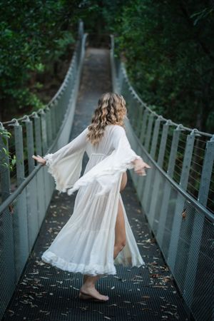 A flowing white dress twirls on a suspension bridge surrounded by lush greenery.