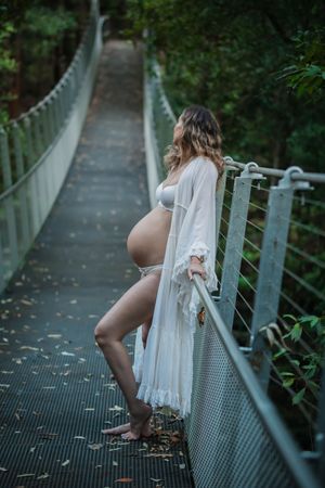 A figure in white lace stands on a metal walkway surrounded by lush greenery in a moody forest setting.