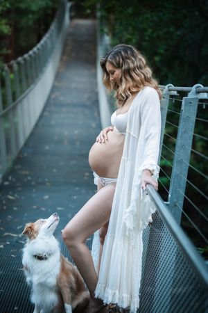 Maternity photoshoot on a bridge with a white flowing dress and small dog looking up at expectant mother.