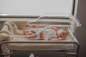 Newborn baby wearing white sleeps peacefully in hospital bassinet with medical monitoring equipment nearby.