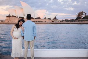 Couple in white and blue attire walking along beach at sunset with ocean and Sydney Opera House in background.