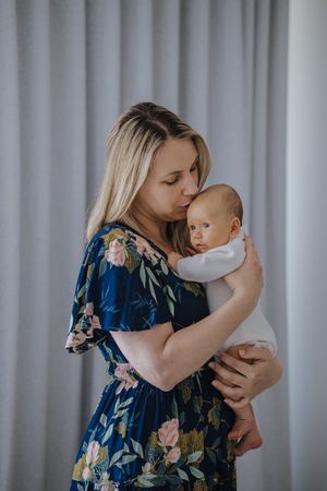 Mother in floral blue dress lovingly holds and kisses baby in tender moment against light curtain background.
