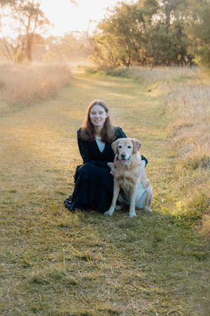 A young woman in a dark dress sits with a golden Labrador on a grassy path at golden hour.