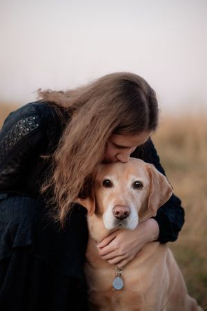 A young woman in a black dress tenderly kisses her golden Labrador dog outdoors in a field.